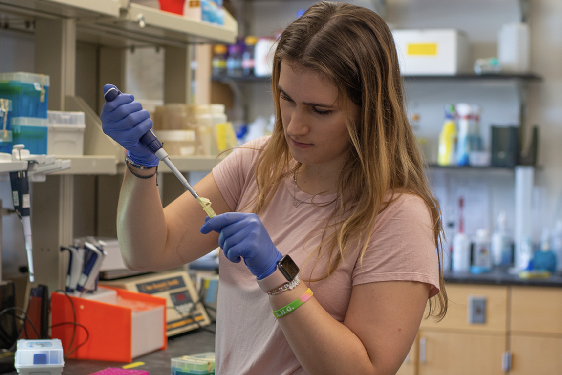 A college student with long hair wearing blue gloves uses a dropper in a biochemistry lab